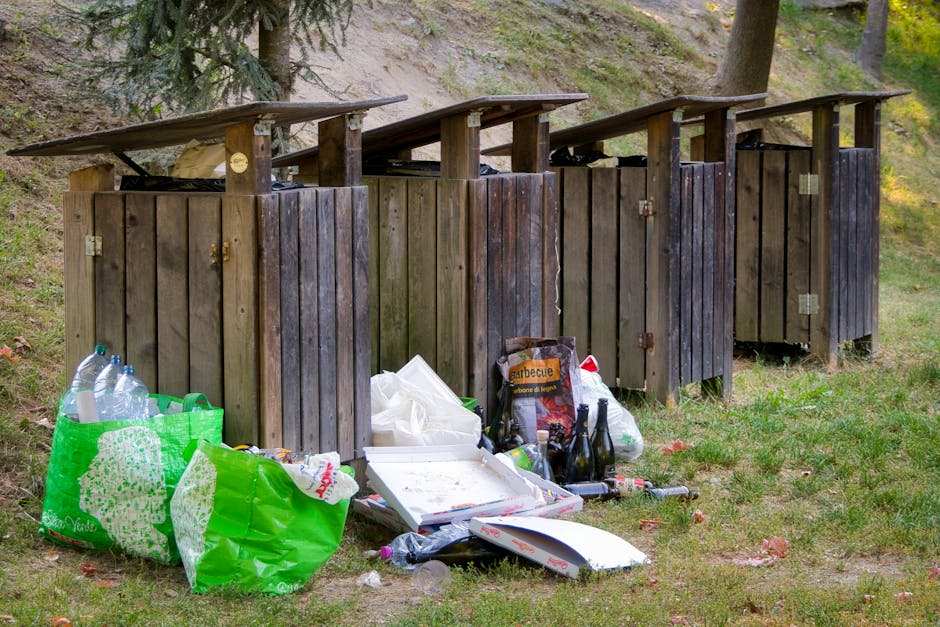 A clear view of a large pile of assorted waste materials situated on a paved driveway or outdoor area. The waste includes various items such as cardboard boxes, plastic containers, metal scrap, and wo