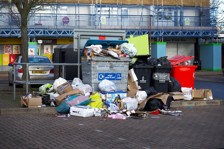 A collection of mixed waste including black plastic garbage bags, a yellow plastic container, and a dirty, stained car seat discarded on an asphalt surface in an outdoor area. The waste is piled again